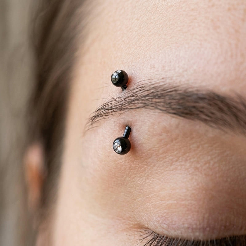 16G black PVD coated steel curved eyebrow barbell with clear CZ gem ball ends, worn in an eyebrow piercing.